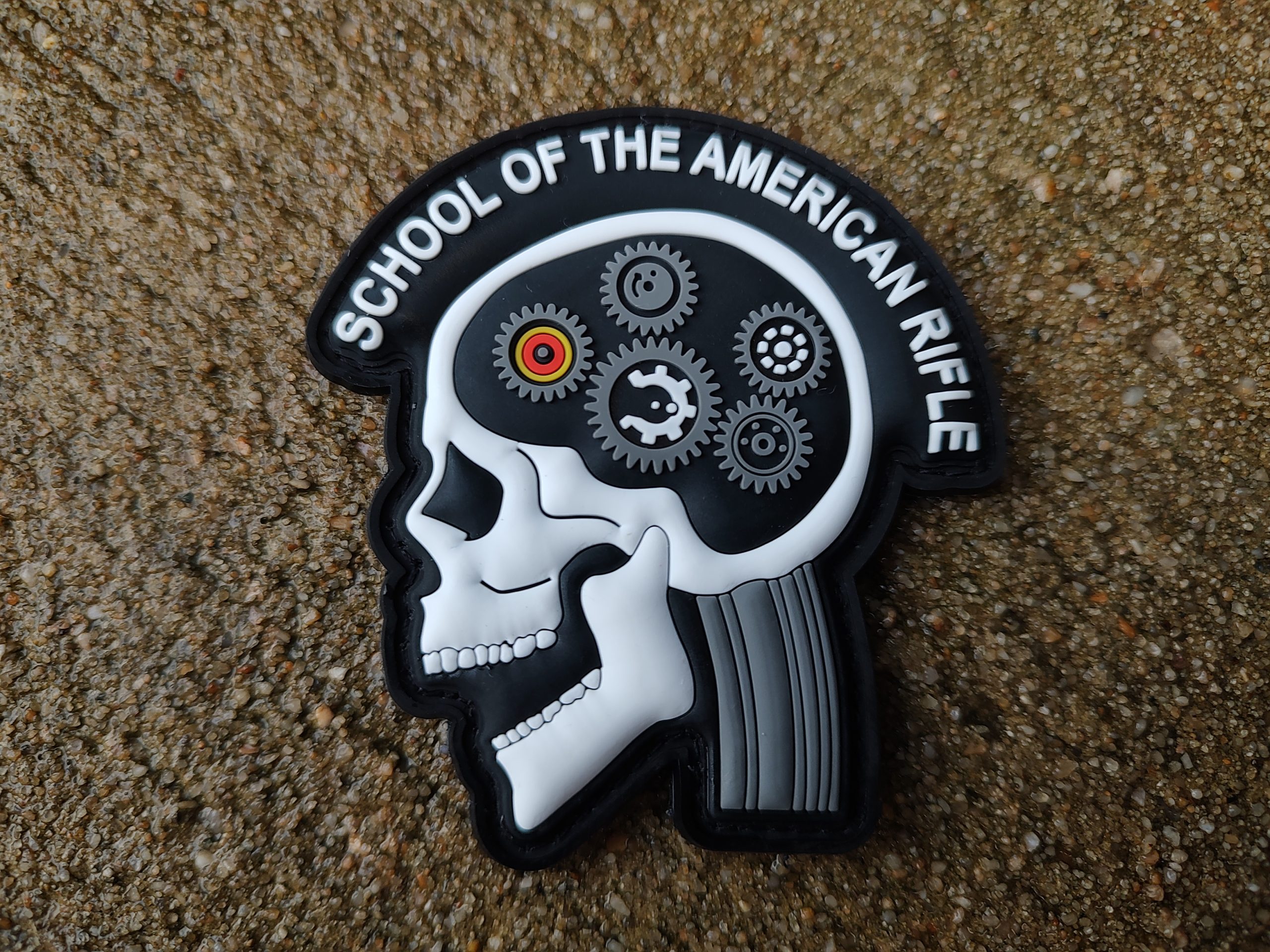 A "SOTAR PVC Patch - MK2 White," featuring a side profile of a skeleton's head filled with gears and labeled "School of the American Rifle," placed on a rough textured surface.