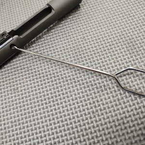 The SOTAR TFPRPRT - MK3 "Tactical Firing Pin Retaining Pin Removal Tool", with its looped handle, is inserted into a slot on a black mechanical component, placed on a gray textured surface.