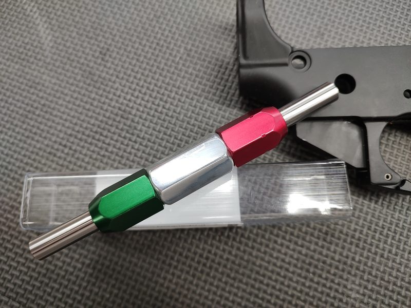 A close-up of the SOTAR AR15/M16 Lower Receiver Selector Bore GO/NO-GO Gauge - MK2, featuring green, silver, and red sections on a hex shaft, placed on a clear plastic box next to a black metal part against a textured background.