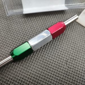 The SOTAR AR15/M16 Lower Receiver Selector Bore GO/NO-GO Gauge - MK2, featuring three hexagonal grips in green, silver, and red, is lying on a textured surface with a clear plastic cover nearby.