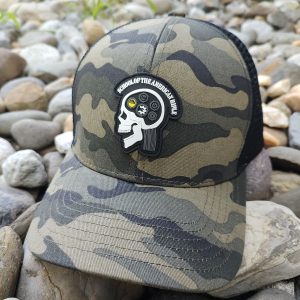 A camouflage baseball cap placed on rocks, featuring a patch with a skull design and the text "School of Thought Nonthinker.