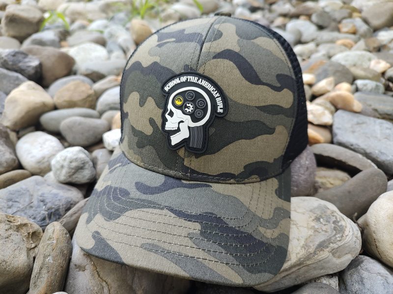 A camouflage baseball cap placed on rocks, featuring a patch with a skull design and the text "School of Thought Nonthinker.