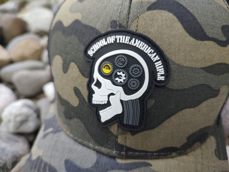 Close-up of a camouflage hat with a skull patch. The patch, labeled "School of the American Rifle," depicts a skull with visible gears inside the head.