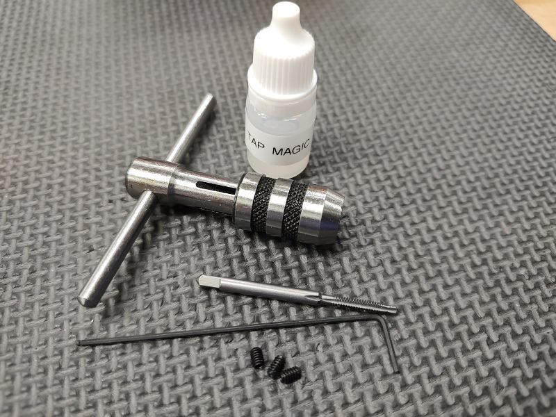 A small bottle labeled 'AR15 Lower Receiver - Captured Takedown Spring - Tap Kit,' a T-handle tap wrench, a tap, two miniature screws, and a fine metal rod are placed on a textured surface.