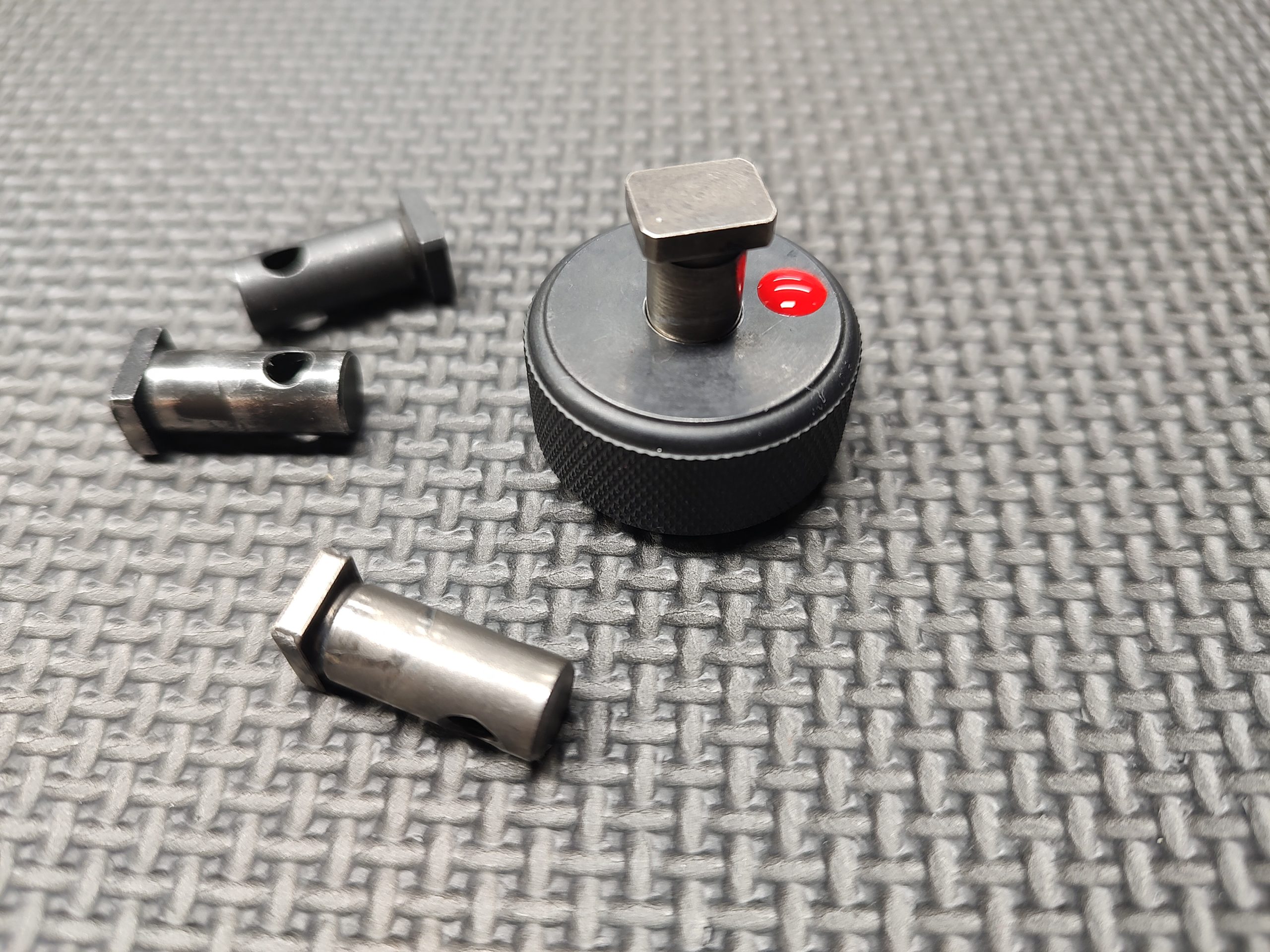 The SOTAR AR15 Cam Pin Field Gauge (Reject Gauge Only), a black cylindrical tool featuring a red button, is positioned on a textured surface alongside three metallic components with holes.