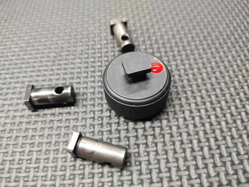 A circular black knob alongside three metallic cylindrical pieces rests on a textured surface, resembling the SOTAR AR15 Cam Pin Field Gauge (Reject Gauge Only).