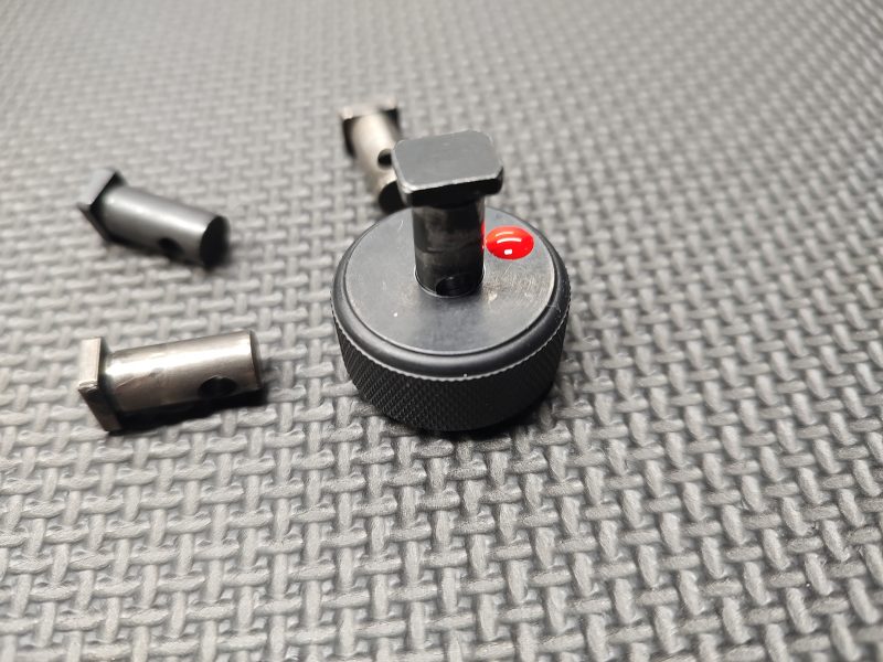 Detailed view of the SOTAR AR15 Cam Pin Field Gauge (Reject Gauge Only), showcasing its black metal design with a threaded knob and red accent, resting on a textured surface with various cylindrical metal components around it.