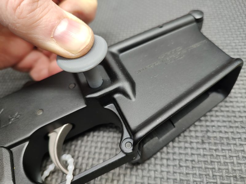 Close-up of a person's hand using the Magazine Button Install Tool on a firearm’s magazine well on a textured surface.