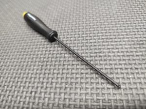 A Magazine Button Install Tool with a black handle and long, thin shaft rests on a textured gray surface.