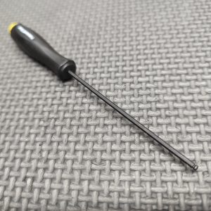 A Magazine Button Install Tool with a black handle and long, thin shaft rests on a textured gray surface.