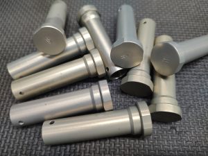 A pile of cylindrical metal rods with flat ends and circular grooves, lying on a textured dark surface.