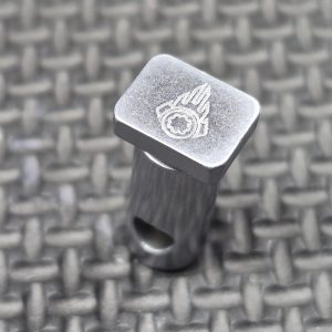 The AR15/M4 Milspec Hard Chrome Cam Pin by Obsidian Arms, featuring a rectangular metallic design with an engraved logo on top, stands upright on a textured, interlocking surface.